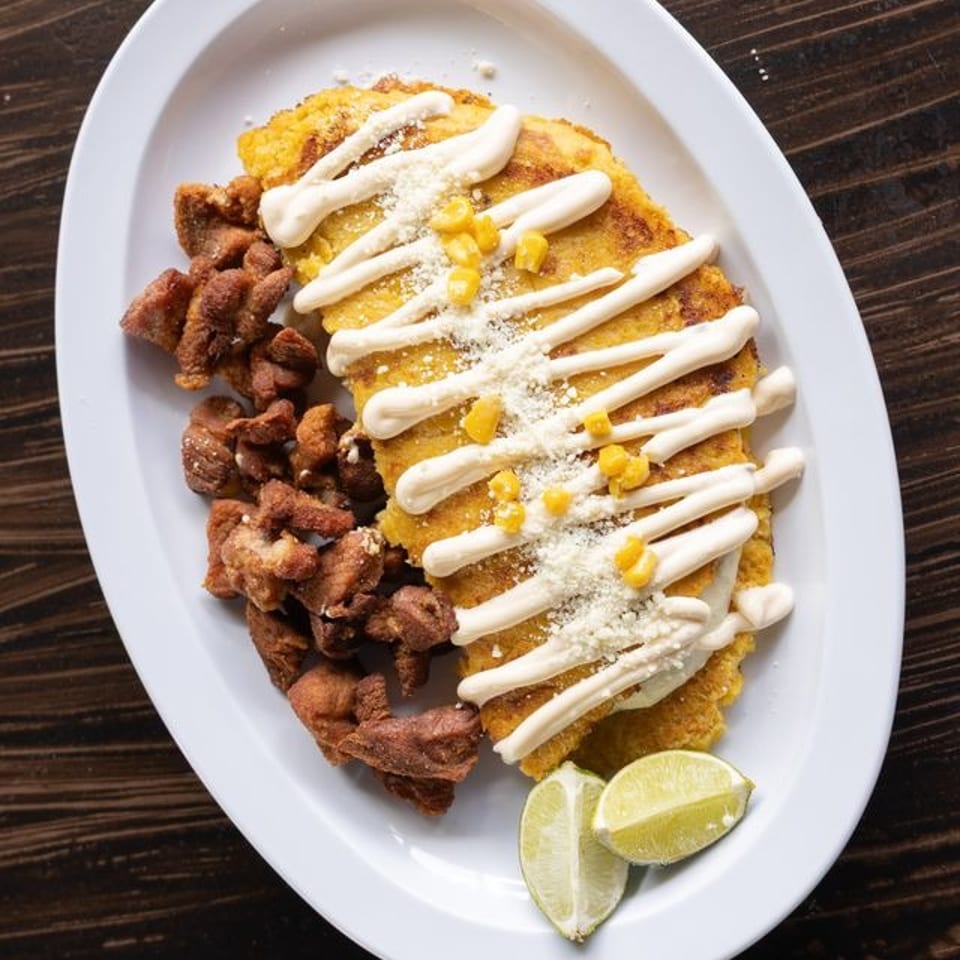 Breaded corn-topped entrée drizzled with cream sauce, served with crispy fried meat and lime wedges on a white platter.
