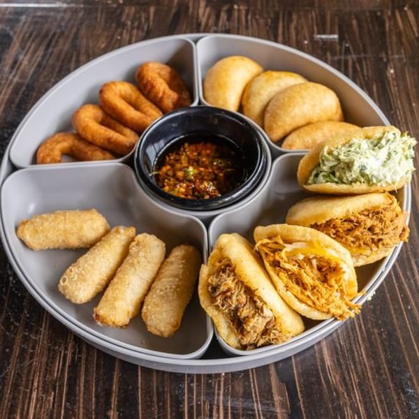 Round serving platter holds assorted fried pastries, stuffed sandwiches, spring rolls, and dipping sauce on a wooden table.