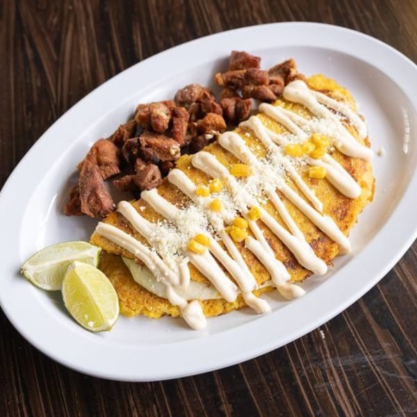 Golden corn cakes topped with grilled meat, corn, crema, and cheese, served with lime wedges on a white plate.