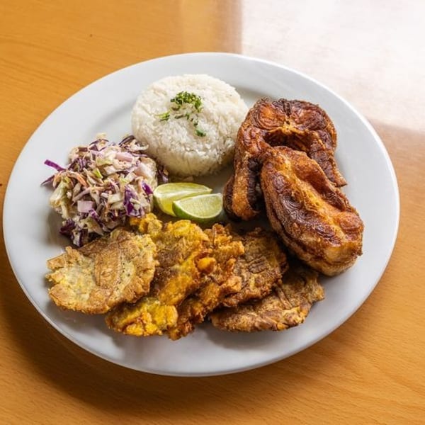 Plate of fried plantains, crispy fried entrée, rice, cabbage slaw, and lime wedges on a wooden table.