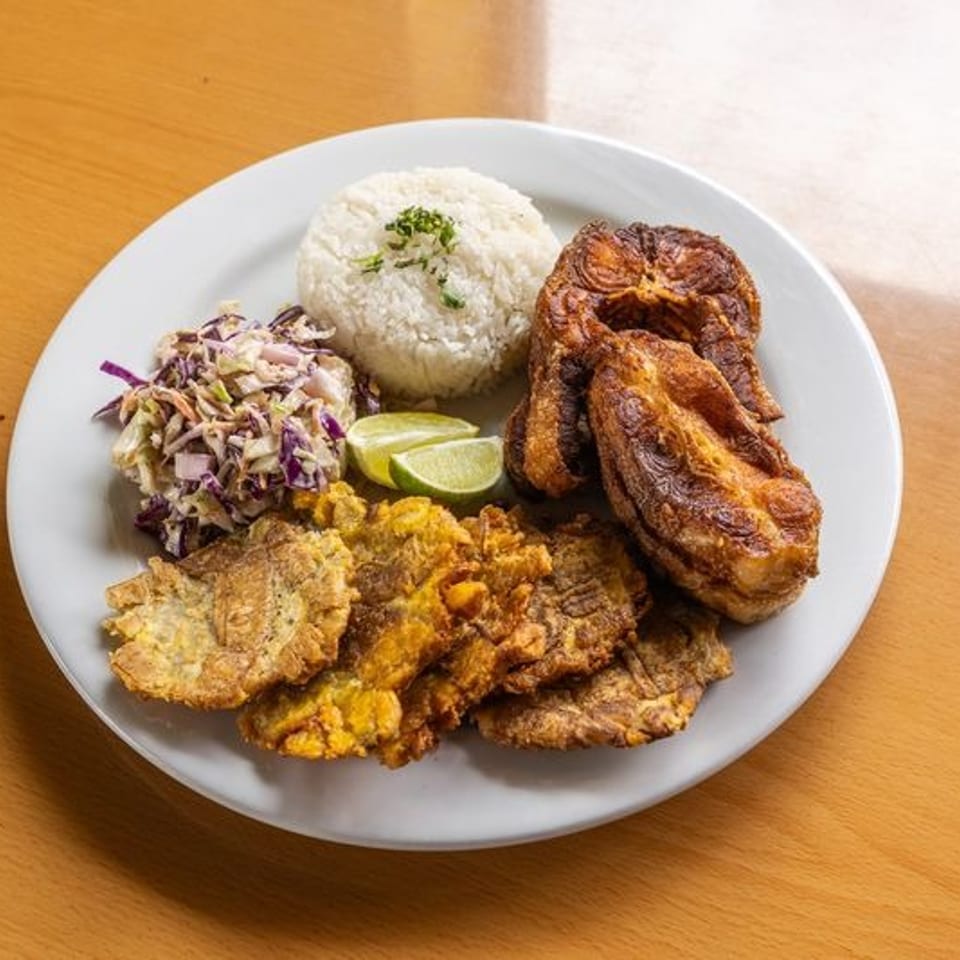 Plate of fried plantains, crispy fried entrée, rice, cabbage slaw, and lime wedges on a wooden table.