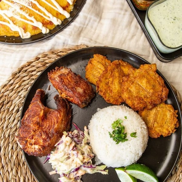 Overhead spread of crispy fried chicken, plantains, white rice, coleslaw, lime wedges, sauces, and golden fried appetizers on black plates.