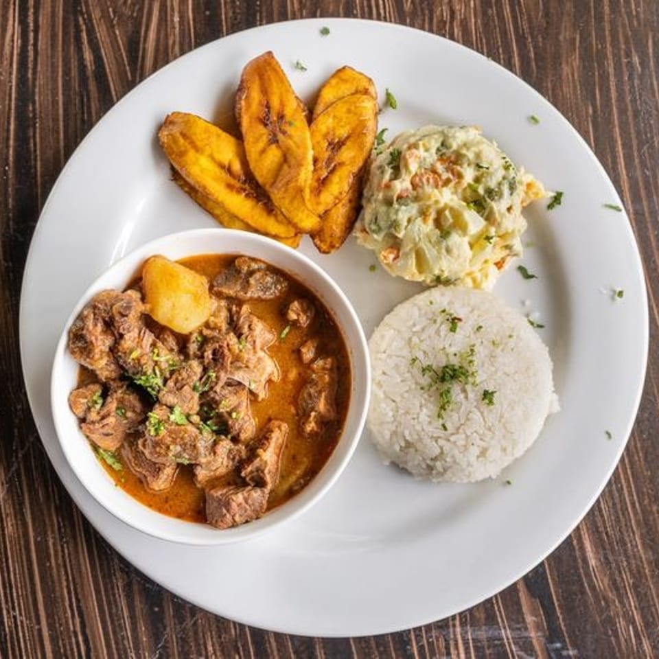 Plate of braised meat stew, white rice, fried plantains, and creamy vegetable salad on a wooden table.