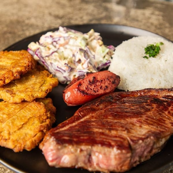 Black plate featuring grilled steak, sausage, fried plantains, white rice, and creamy coleslaw.