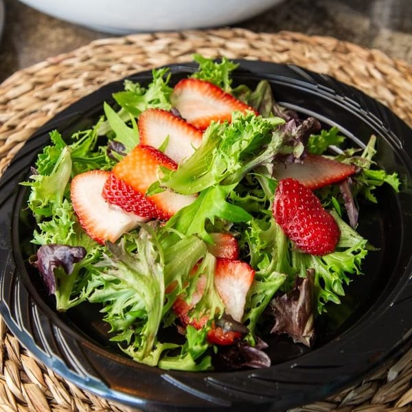 Mixed green salad topped with sliced strawberries, served on a black plate beside a bowl of roasted potatoes and carrots.