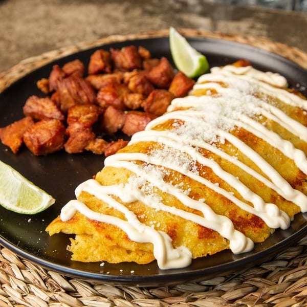 Golden fried tacos topped with cream sauce and cheese, served with seasoned diced meat and lime wedges on a black plate.