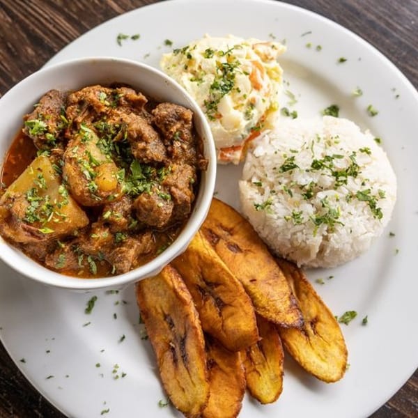 Plate of stewed meat and potatoes with rice, plantains, and creamy salad, garnished with fresh herbs on a wooden table.