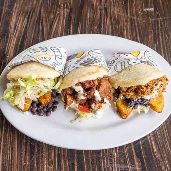 Three stuffed sandwiches with lettuce, beans, fried fillings, sauces, and colorful paper wrappers arranged on a white plate.