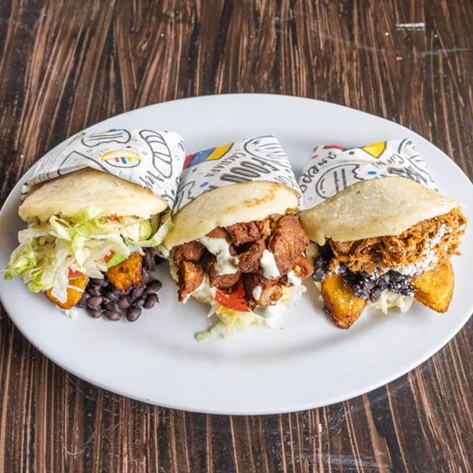 Three stuffed sandwiches with lettuce, beans, fried fillings, sauces, and colorful paper wrappers arranged on a white plate.