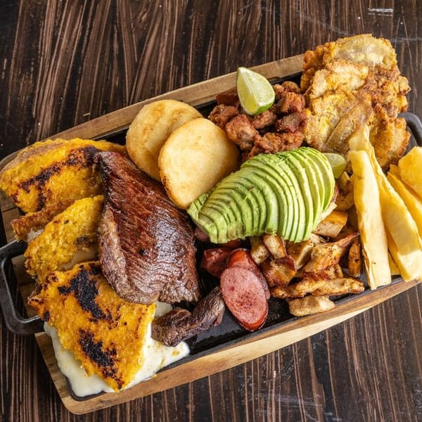 Loaded Latin American grill platter with steak, grilled meats, fried plantains, avocado, potatoes, arepas, and lime on a wooden table.